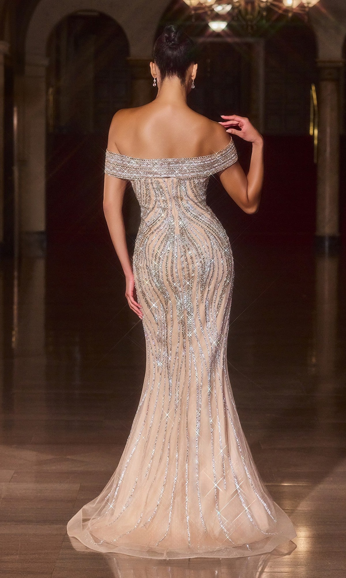 A woman stands on a shiny floor in the Ladivine CD674 long prom dress, an off-the-shoulder, fitted nude sequin gown with silver accents and a subtle mermaid hem, facing away in an elegant, dimly lit interior.